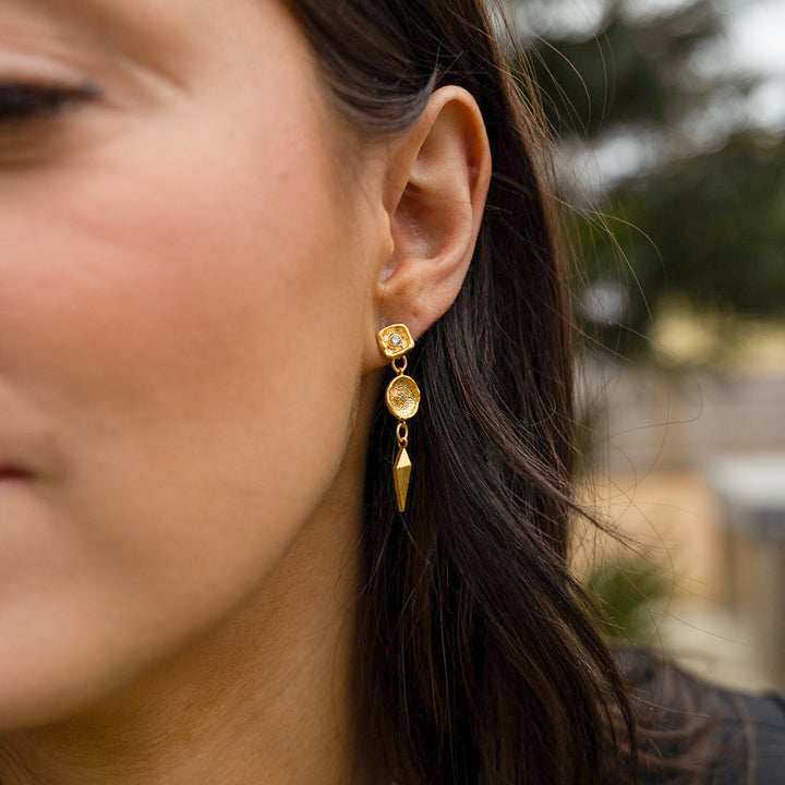 A matte gold dangle earring with a shield charm and a crystal connector. Post earrings with a crystal detail