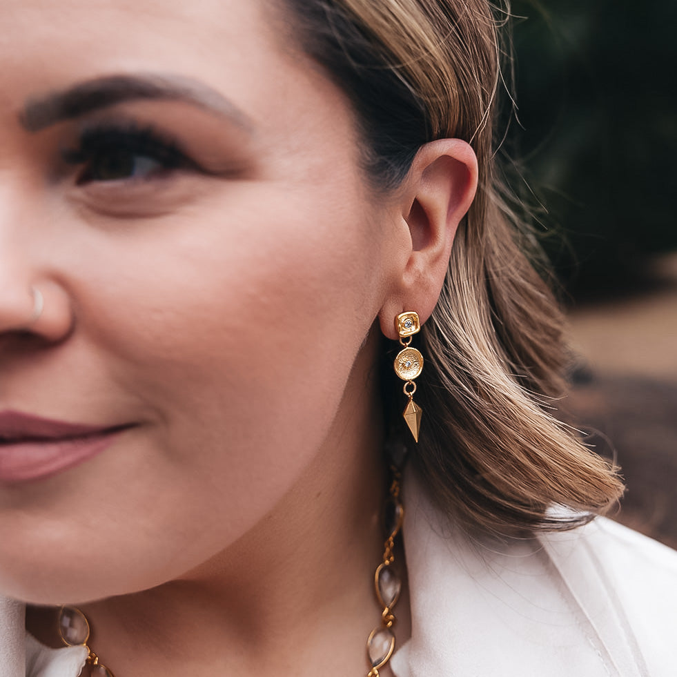 A matte gold dangle earring with a shield charm and a crystal connector. Post earrings with a crystal detail