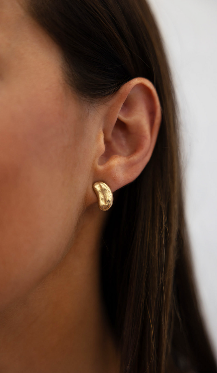 A model wearing A matte gold bean shaped stud earringAn oval hammered dangle earring on oval cutout posts.