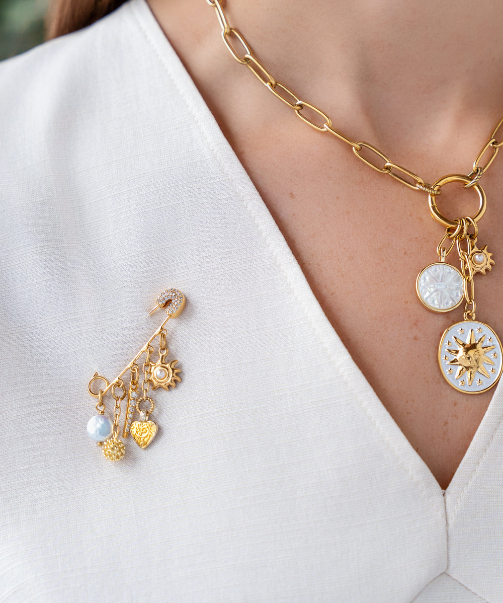 Gold celestial brooch pin with dangling pearl, crystal pavé charm, heart pendant, textured orb, and sunburst charm.