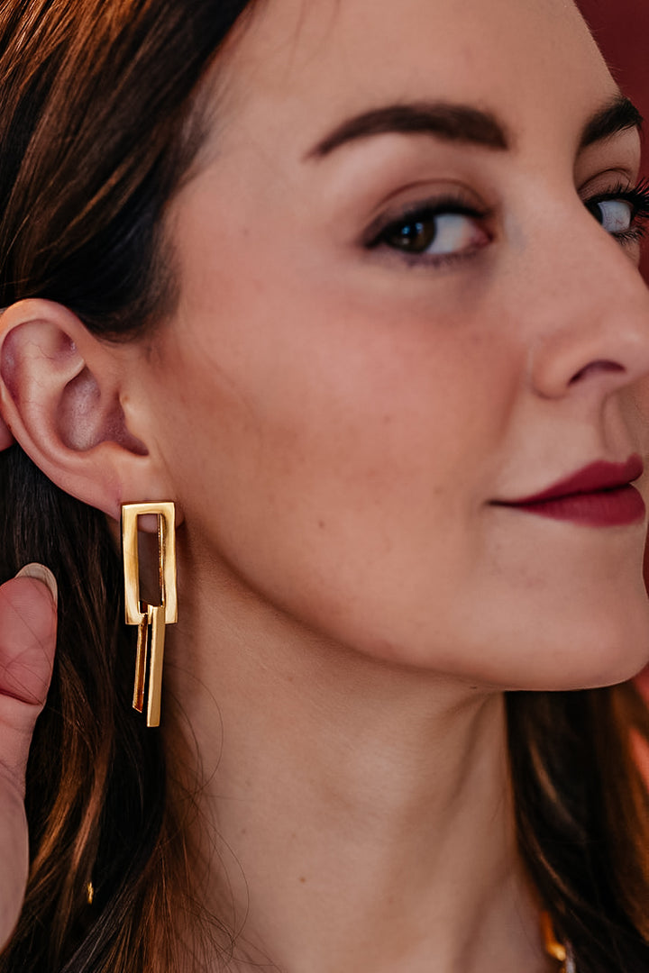 A model wearing A gold dangle earring with interlocking rectangle shapes.